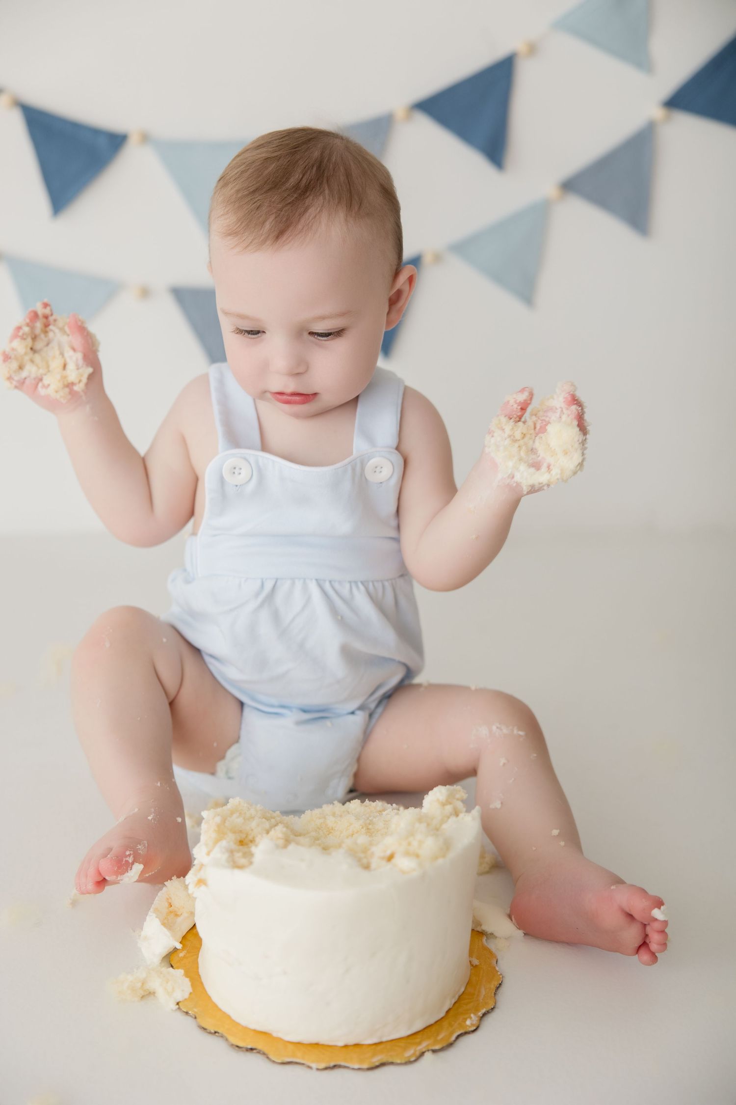 A baby in white romper reaches for and plays with their birthday cake during a cake smash photoshoot.
