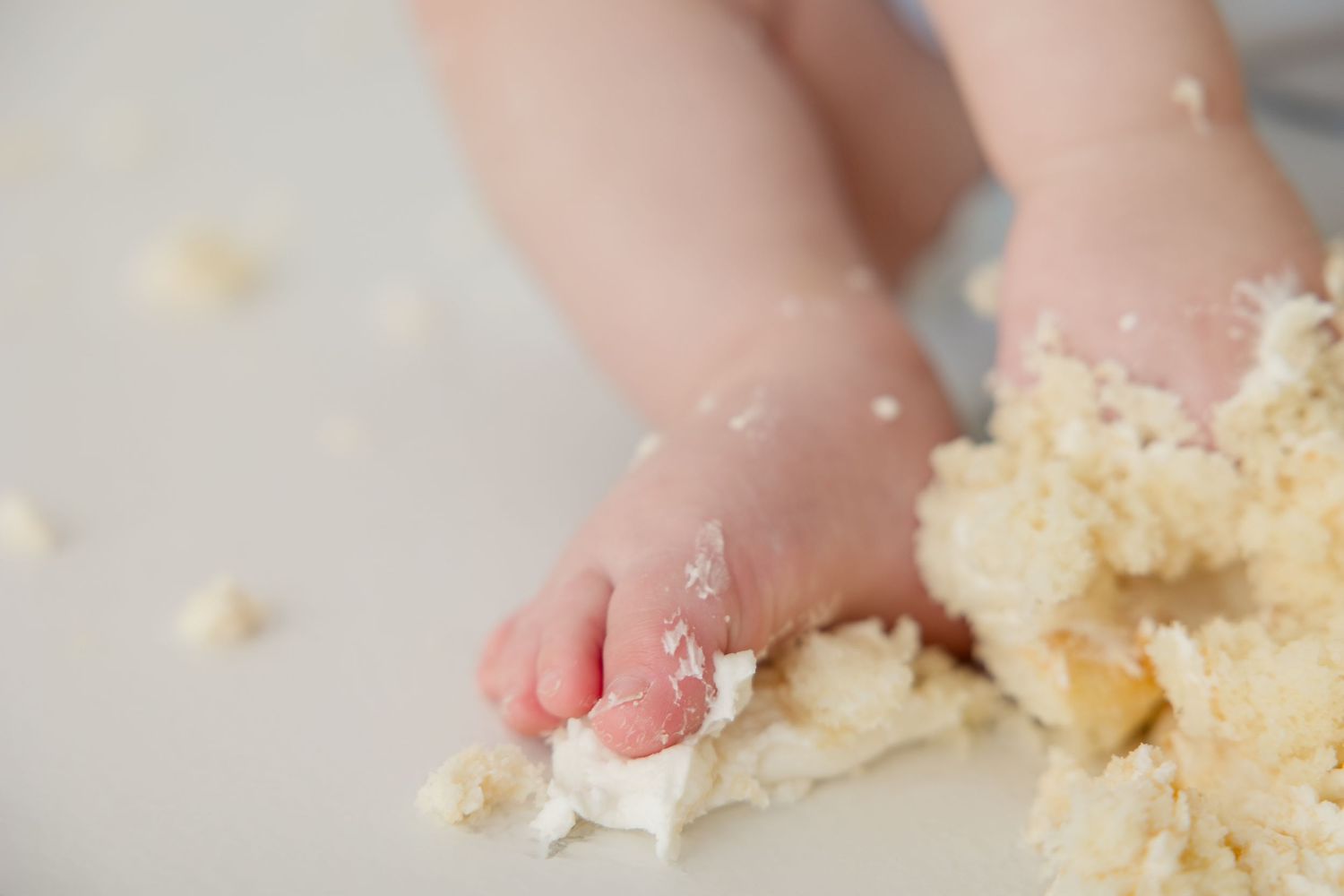 Baby in white outfit messily eating cake with blue bunting flags in background during cake smash photo session.