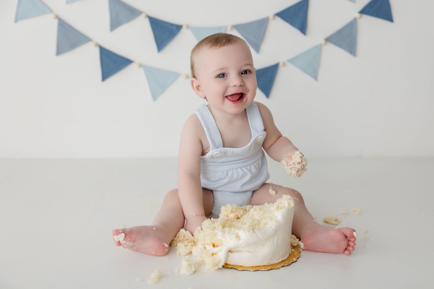 Baby in light blue romper smiles while reaching for birthday cake during first birthday cake smash photo session.
