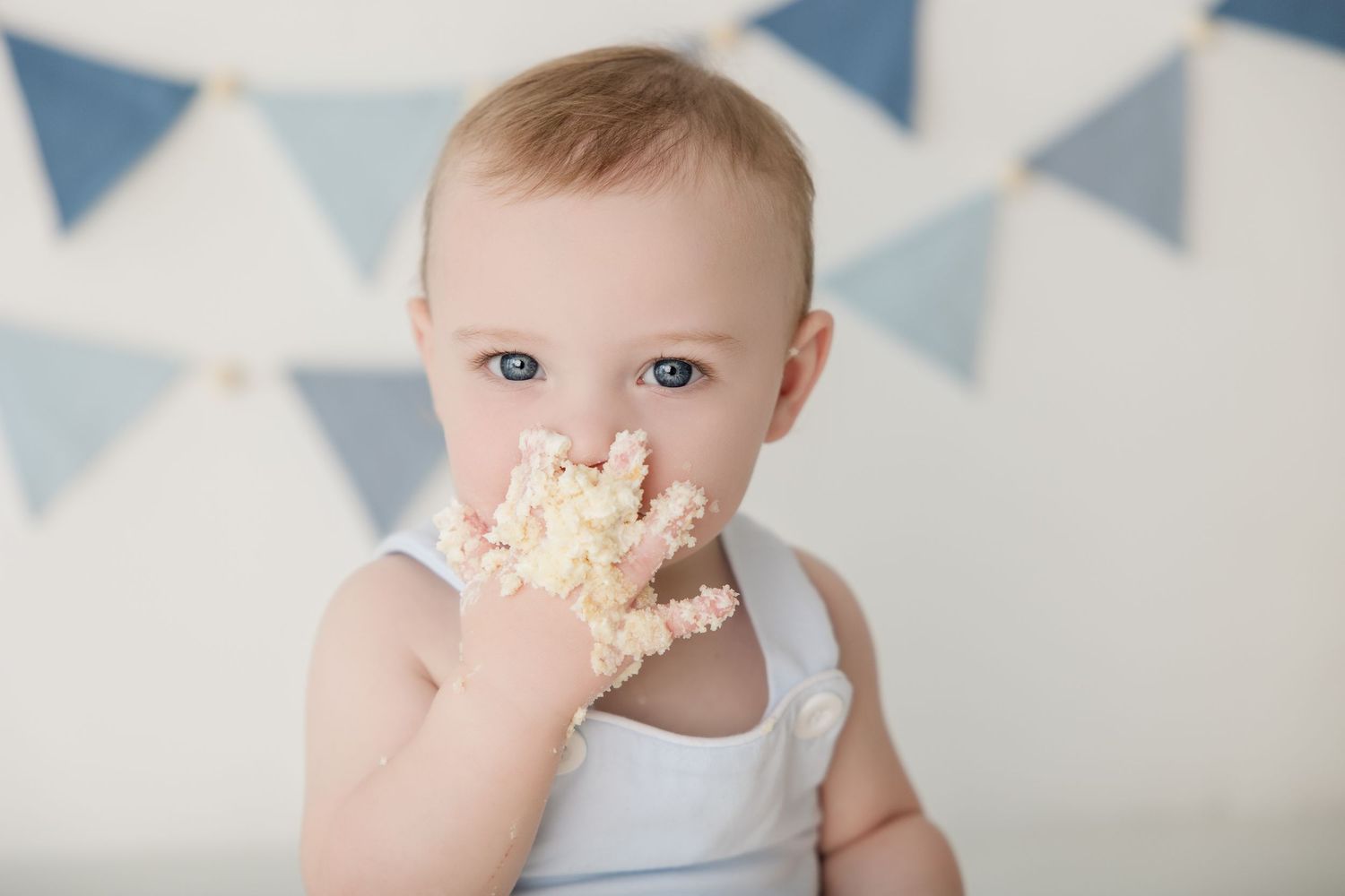 Baby wearing white outfit gets messy while eating birthday cake with blue banner decorations.