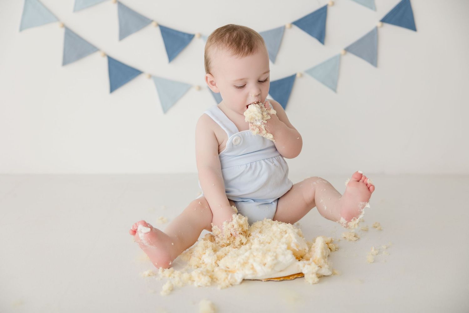 Small baby in blue and white outfit sits among bunting decorations and smashes into a cake during messy cake smash session.
