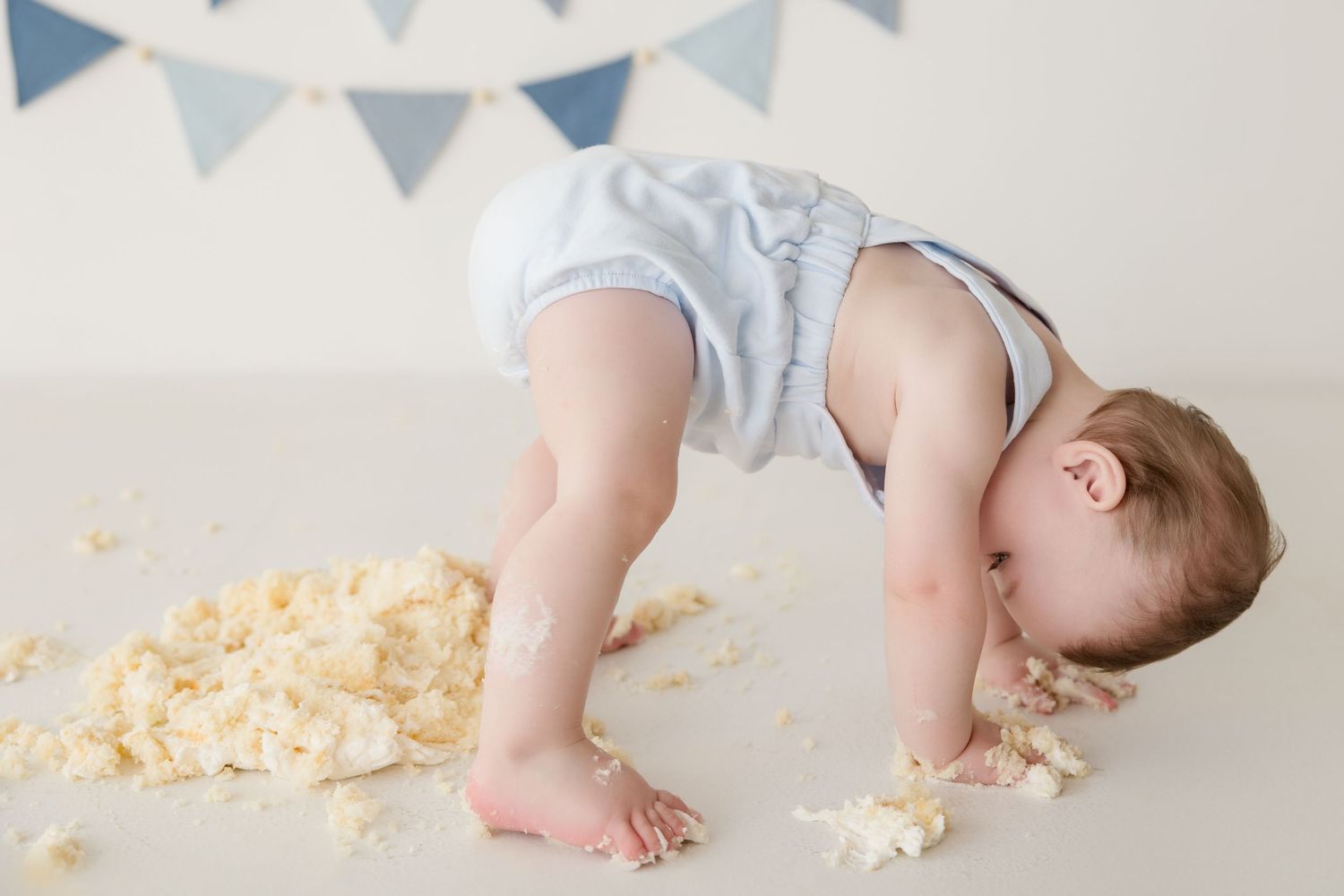 Baby crawling on floor surrounded by smashed cake crumbs with blue triangle banner backdrop.
