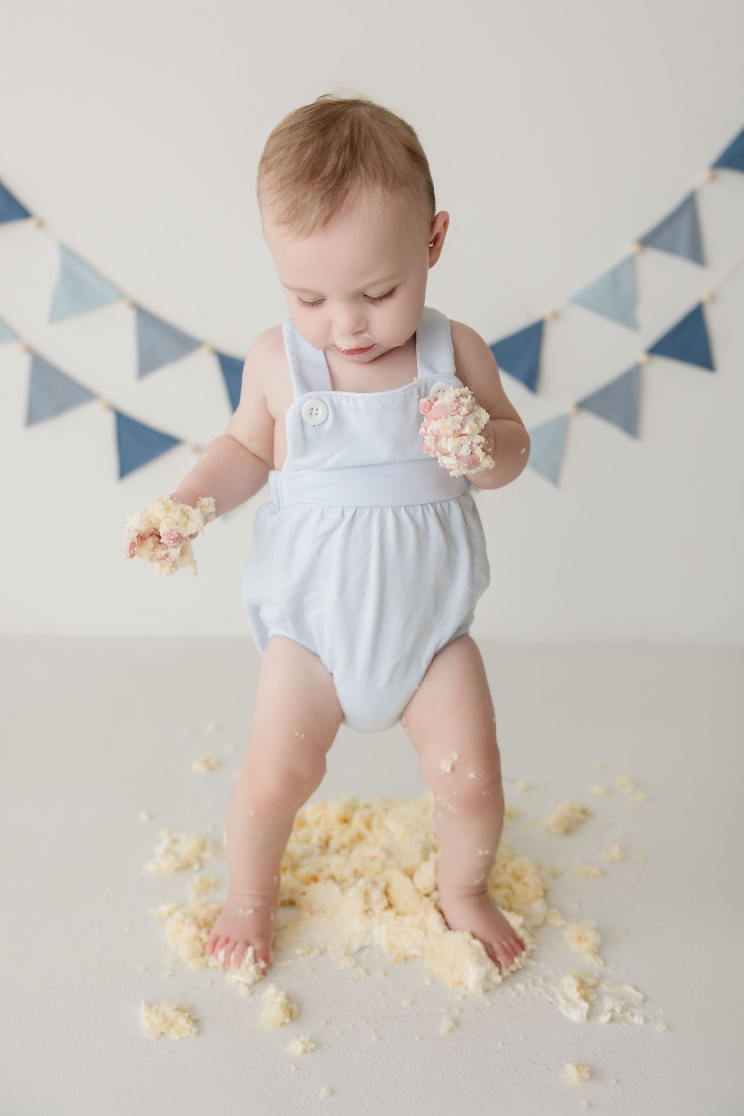 Baby in white romper plays with scattered cake crumbs while blue bunting decorates the background.