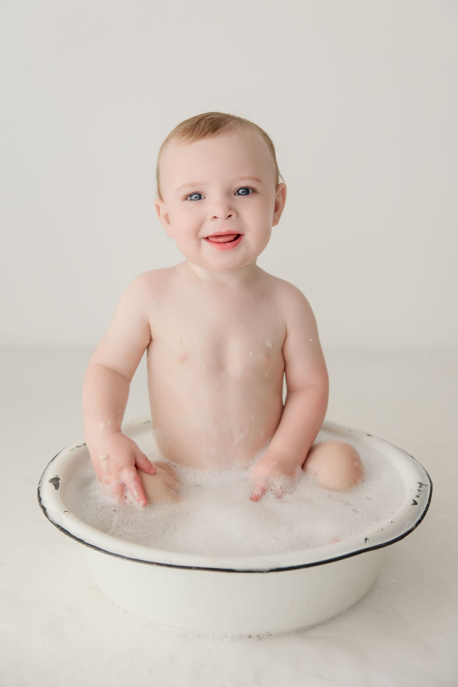 Baby plays in a white bowl filled with suds during a milky bath photography session.