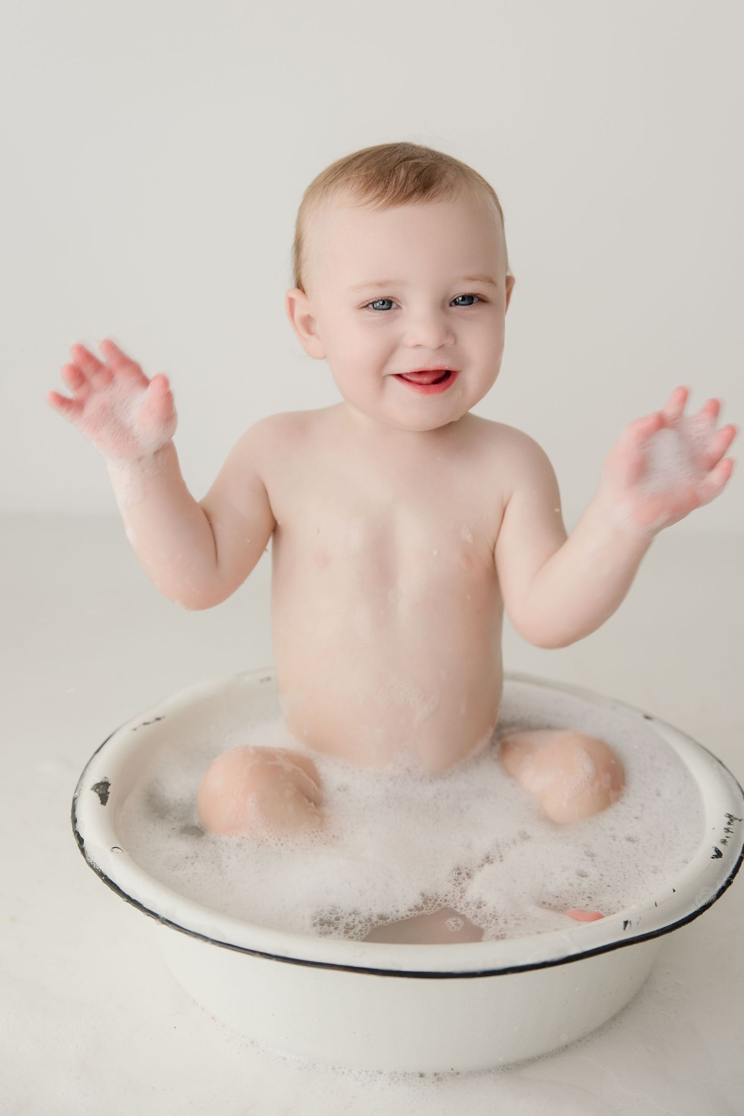 Happy baby splashes in a white basin filled with bubbles during a milk bath photoshoot.