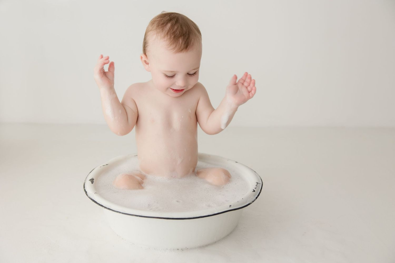 Small baby sits in round white ceramic bowl during minimalist portrait session on light background.