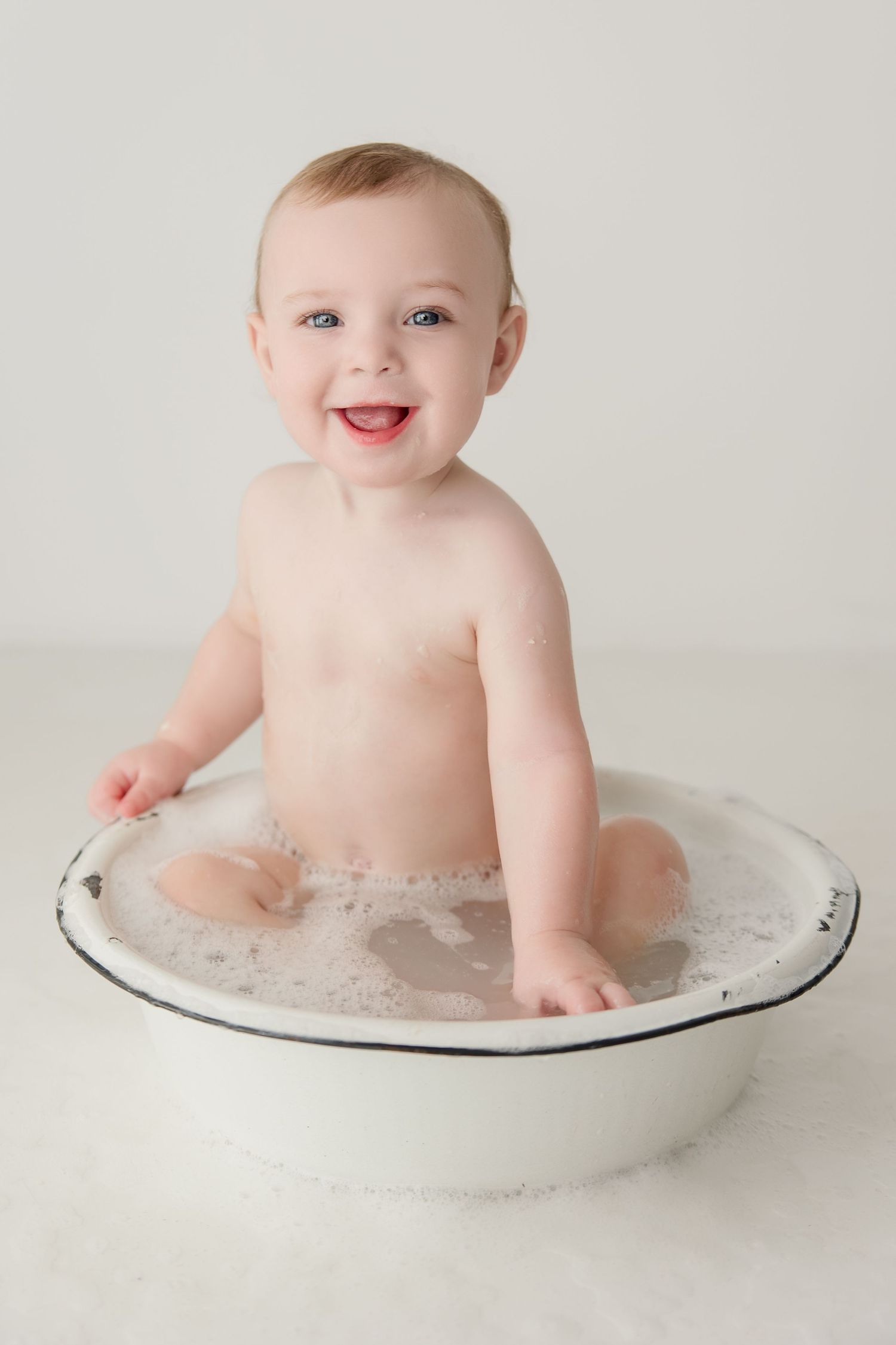 Baby splashes and smiles during bath time in a vintage-style enamel basin against white background.