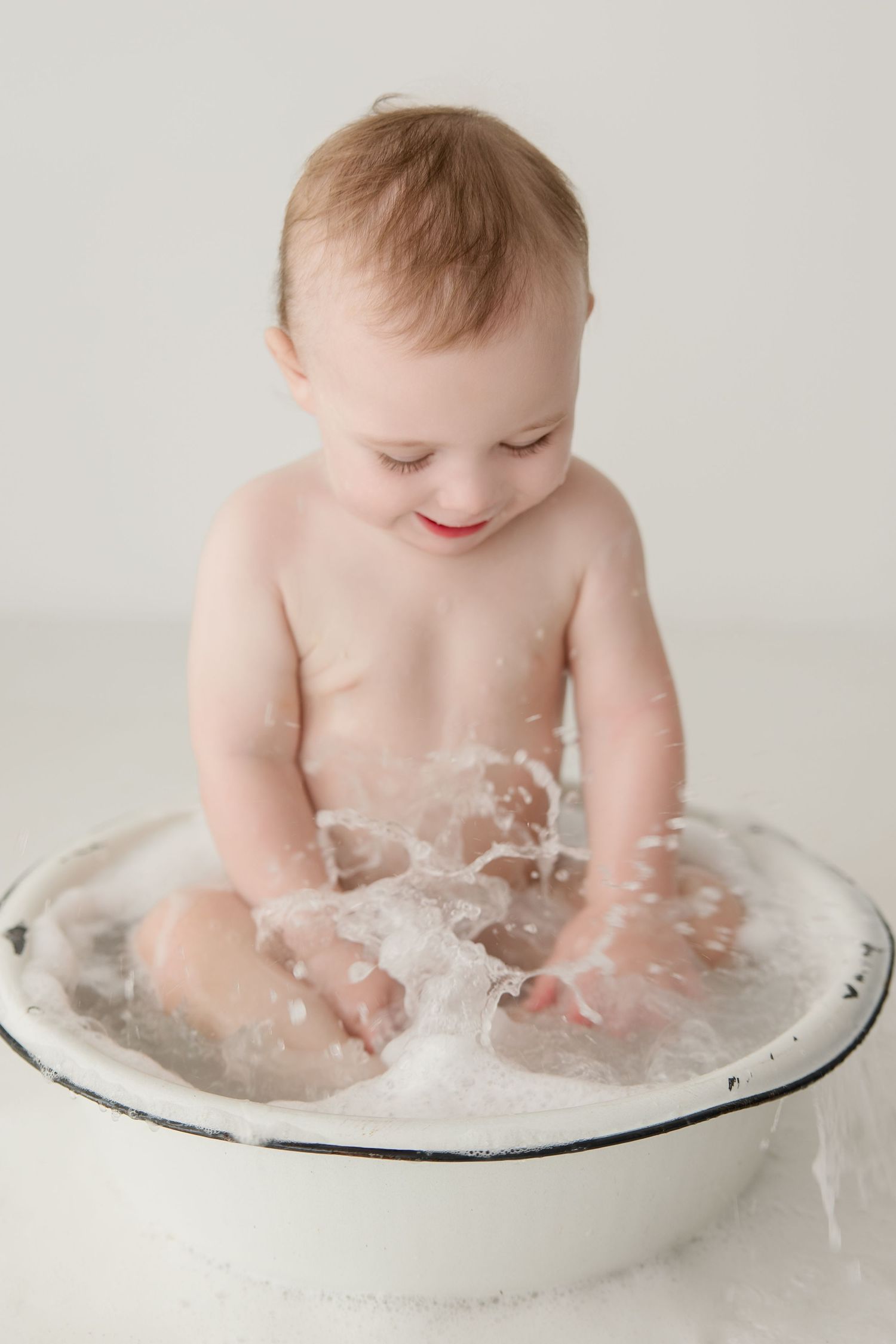 A baby splashes and plays in water during a milk bath photo session with white backdrop.