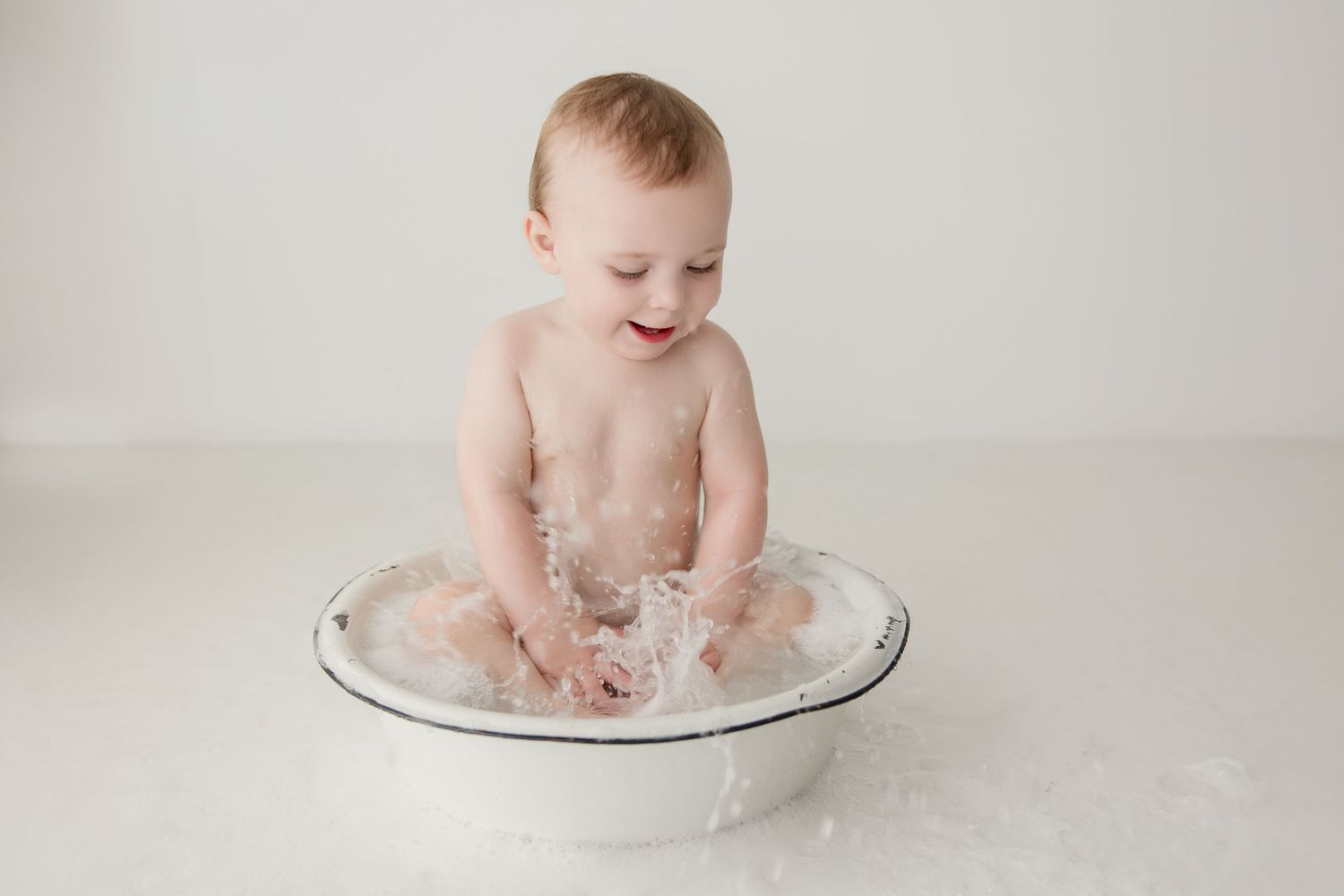 Baby in white sits and plays in a metal bowl during photo session.