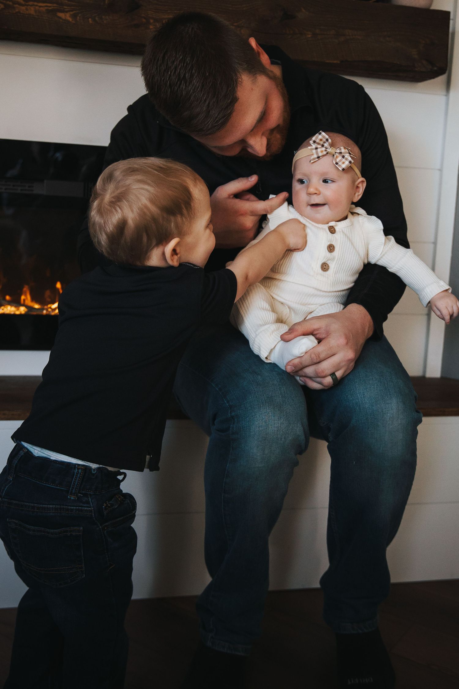 Family moment by fireplace with toddler and baby wearing white outfit.