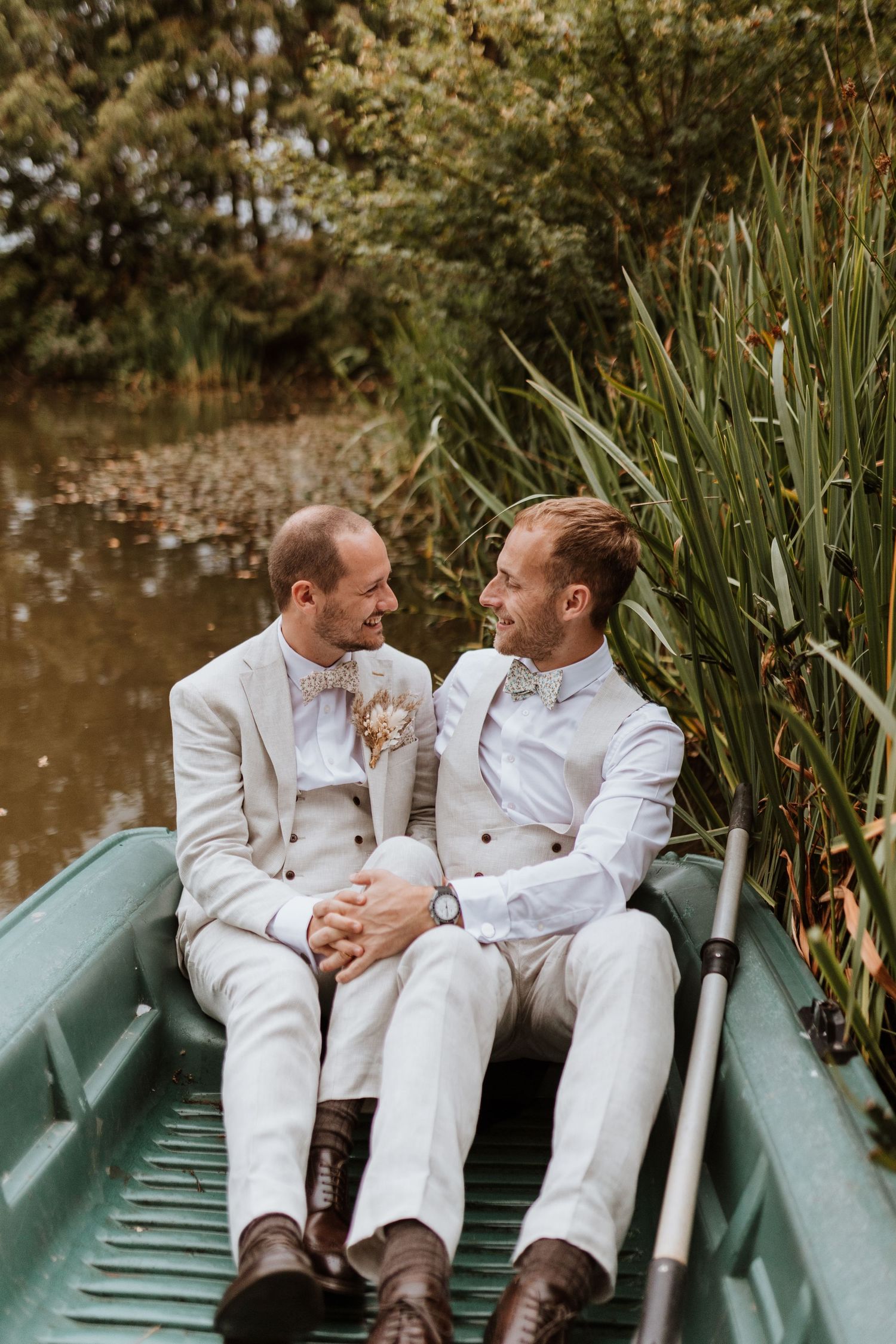 Newly married couple in matching white suits share a joyful moment while sitting in a green rowboat near reeds by a pond at Moat Hall Barns
