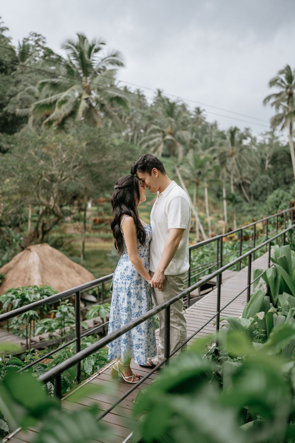 A couple shares a romantic moment on a wooden suspension bridge surrounded by tropical palm trees.