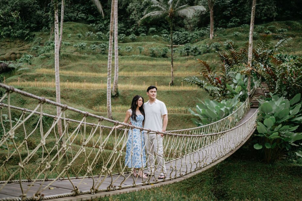 A couple stands together on a suspended rope bridge with palm trees in the background.