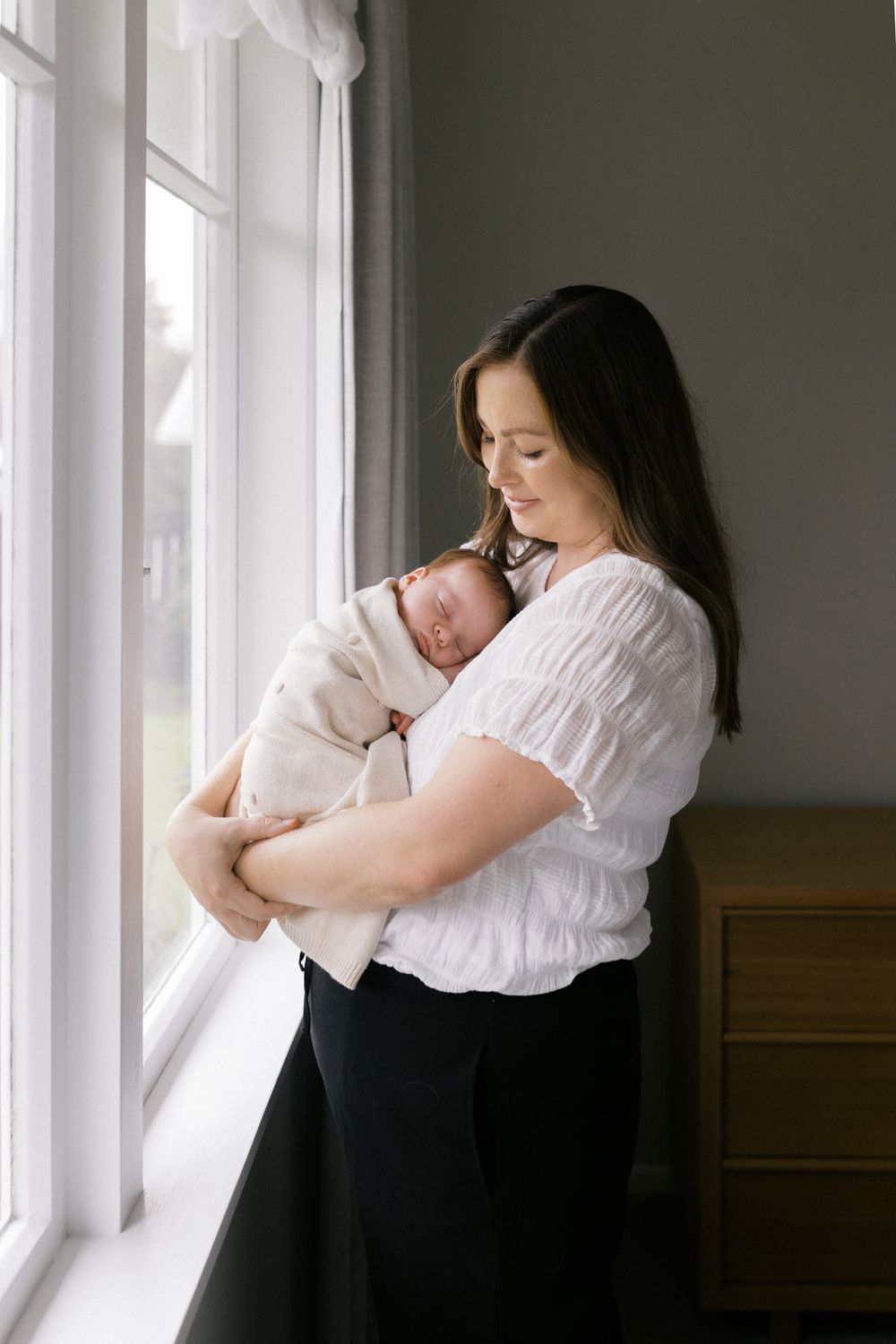 Parent in white top and dark pants by window holding and comforting sleeping baby in tender moment.