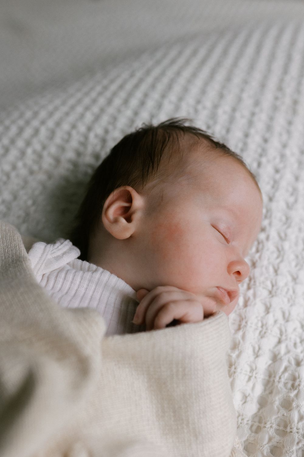 A peaceful newborn sleeping on a textured white knit blanket in soft natural light.