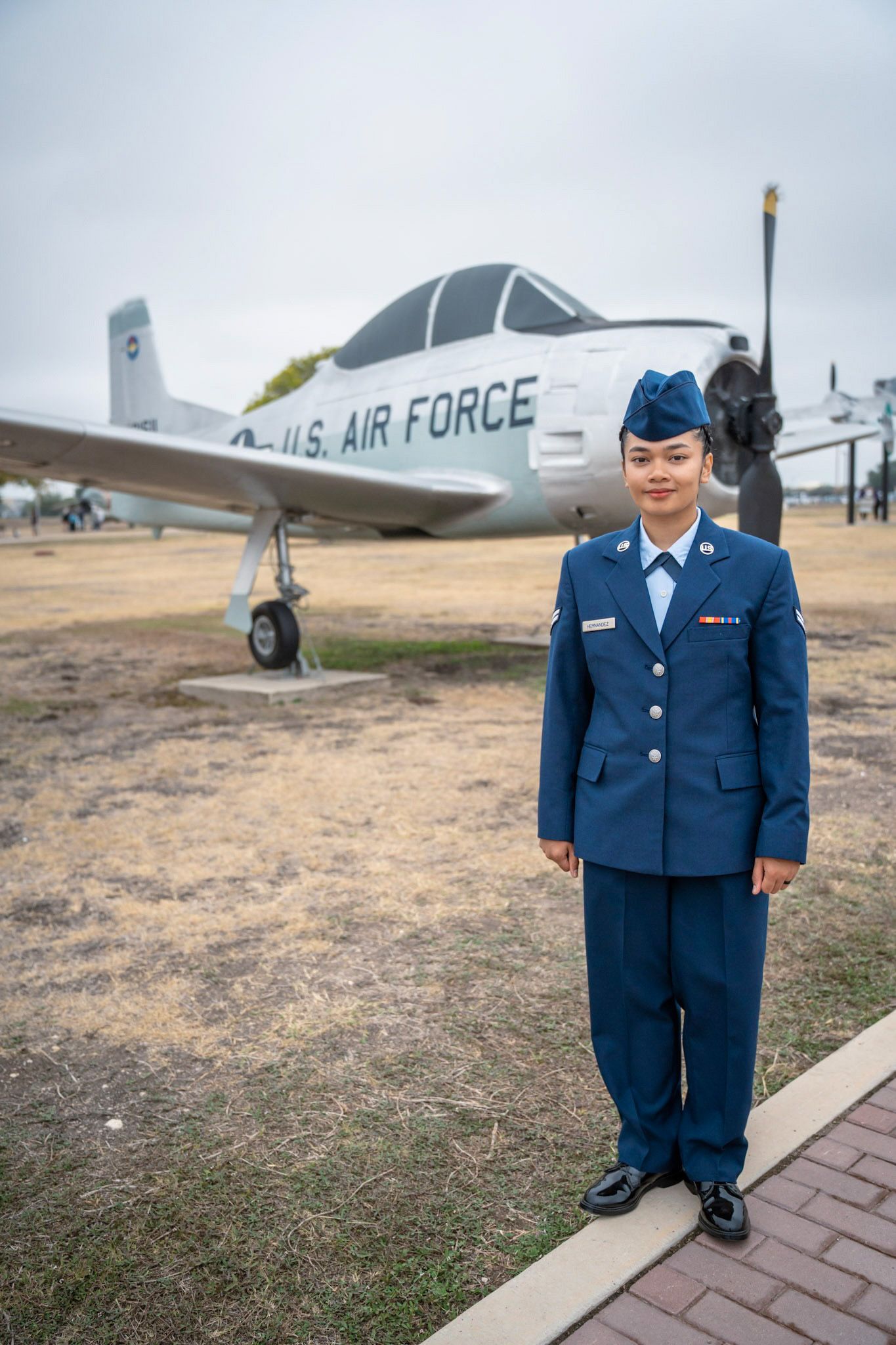 United States Air Force trainee in blue dress uniform posing in front of a vintage aircraft at Lackland AFB.