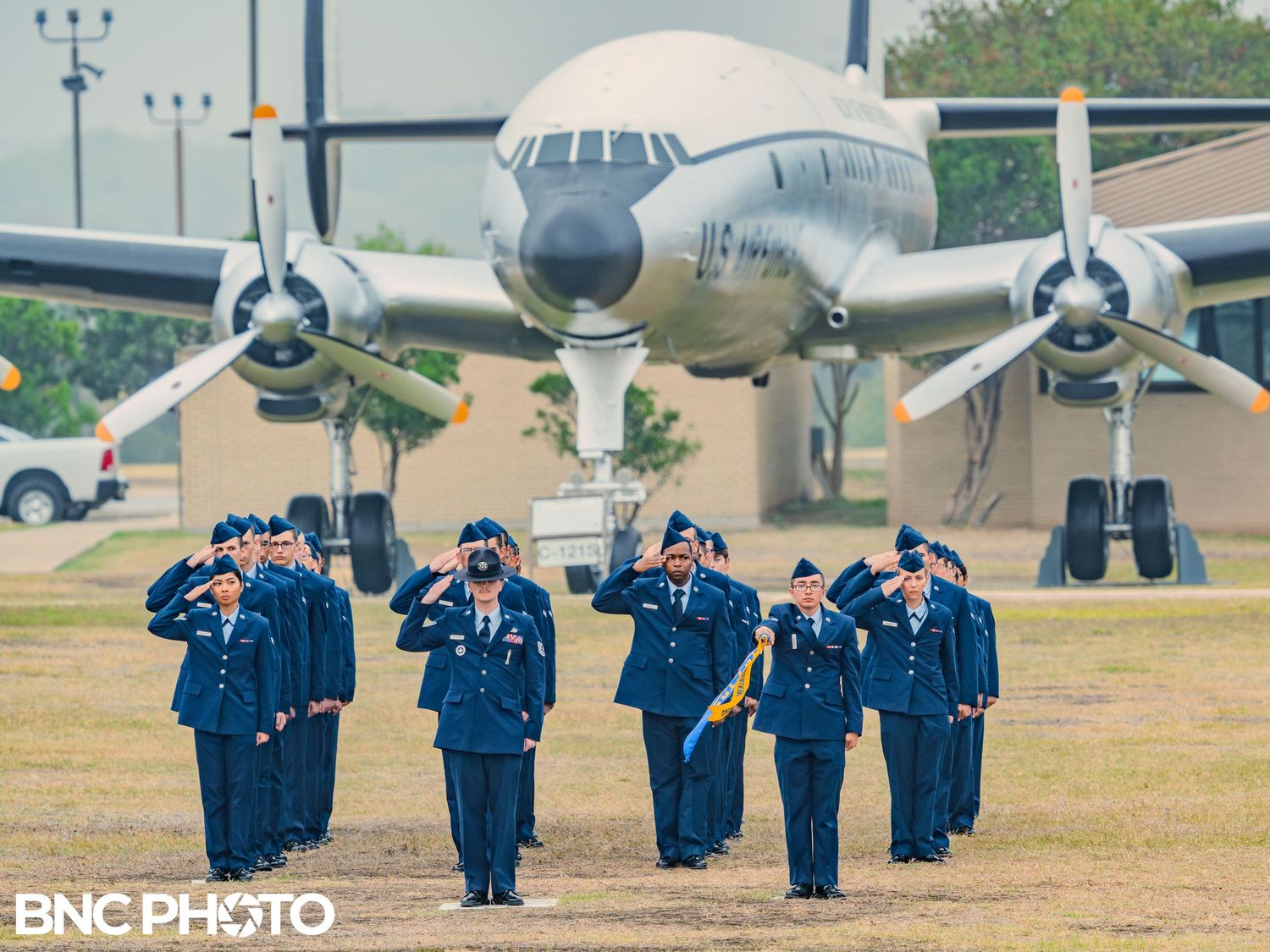 Air Force honor guard saluting and marching on a parade field with a vintage aircraft in the background at Lackland AFB.