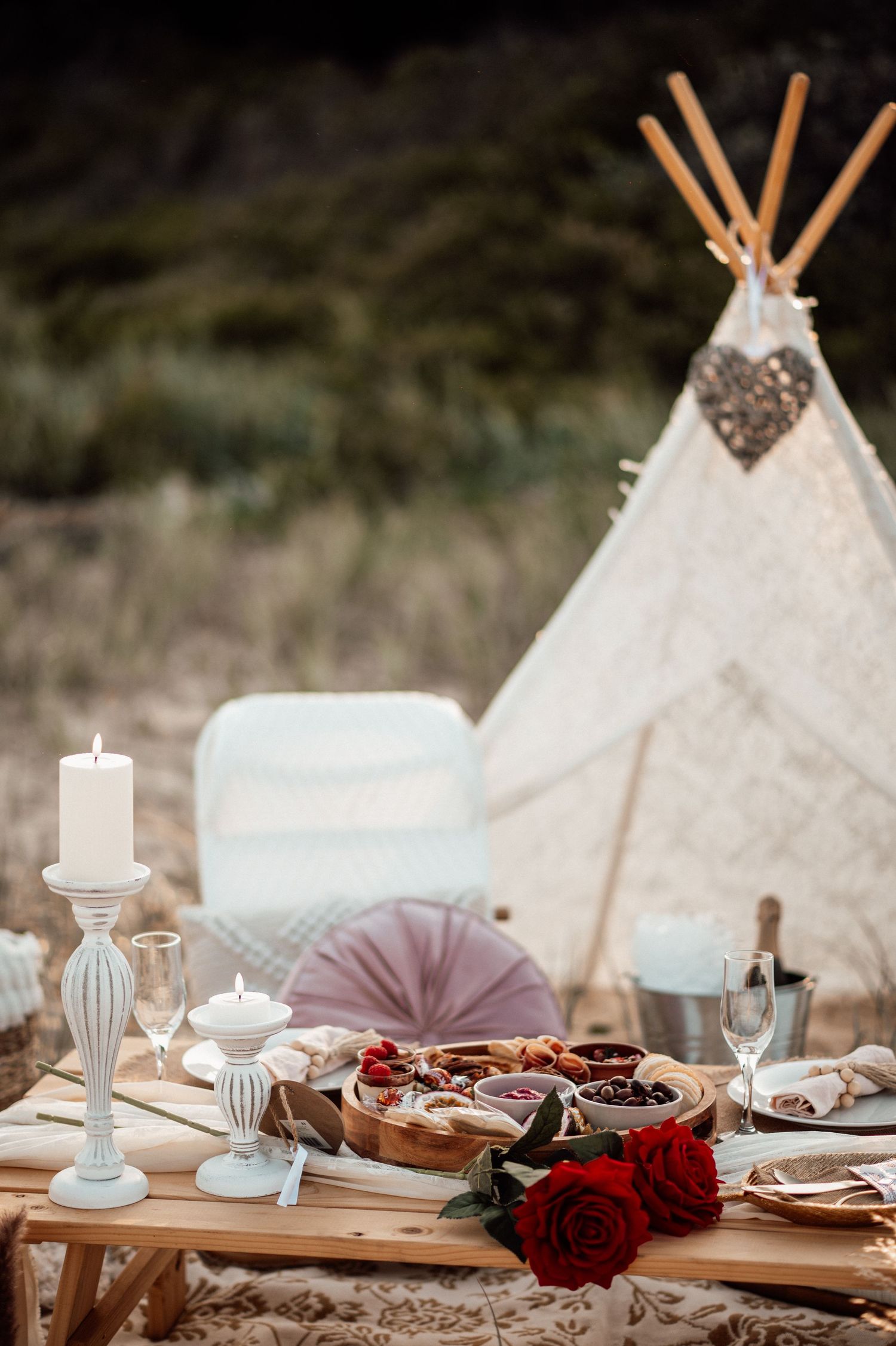 An elegant table setting features white candles, red roses, and vintage decor against a natural outdoor backdrop.