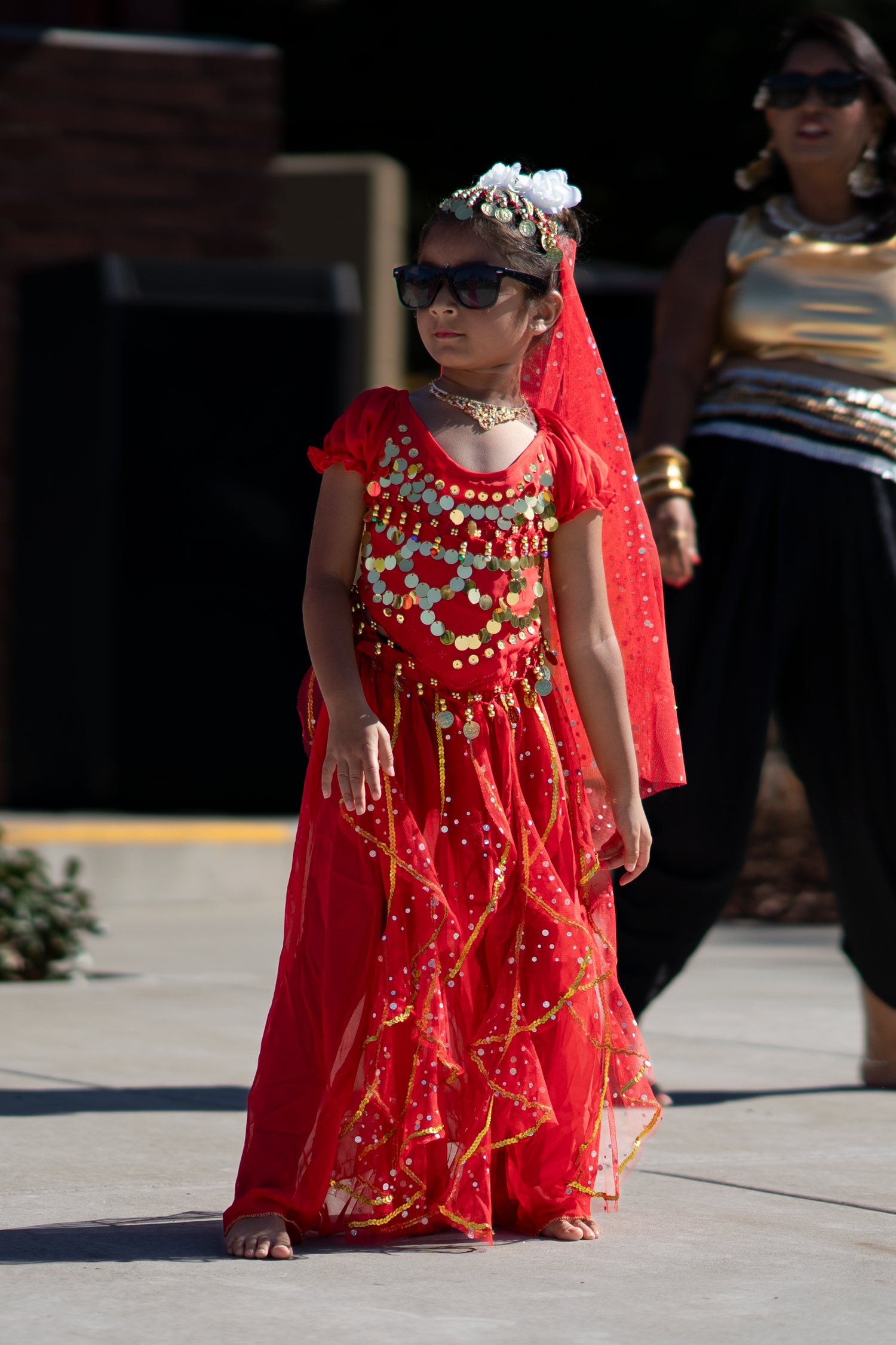 Traditional dancer in ornate red costume with gold jewelry performs on concrete stage.