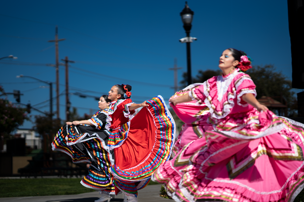 Traditional dancers in pink and red flowing dresses perform against a blue sky backdrop.
