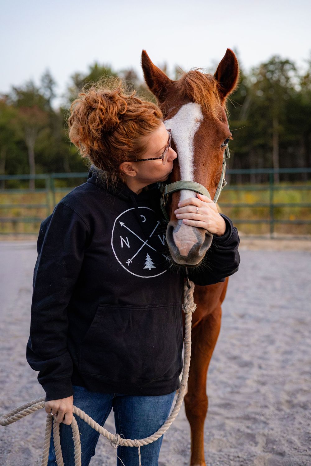 A person in a black hoodie shares a tender moment with a chestnut horse in a sandy riding arena.