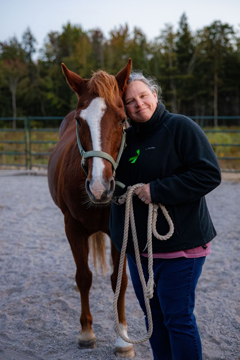 A person in a navy sweater stands next to a brown and white horse in an outdoor riding arena.