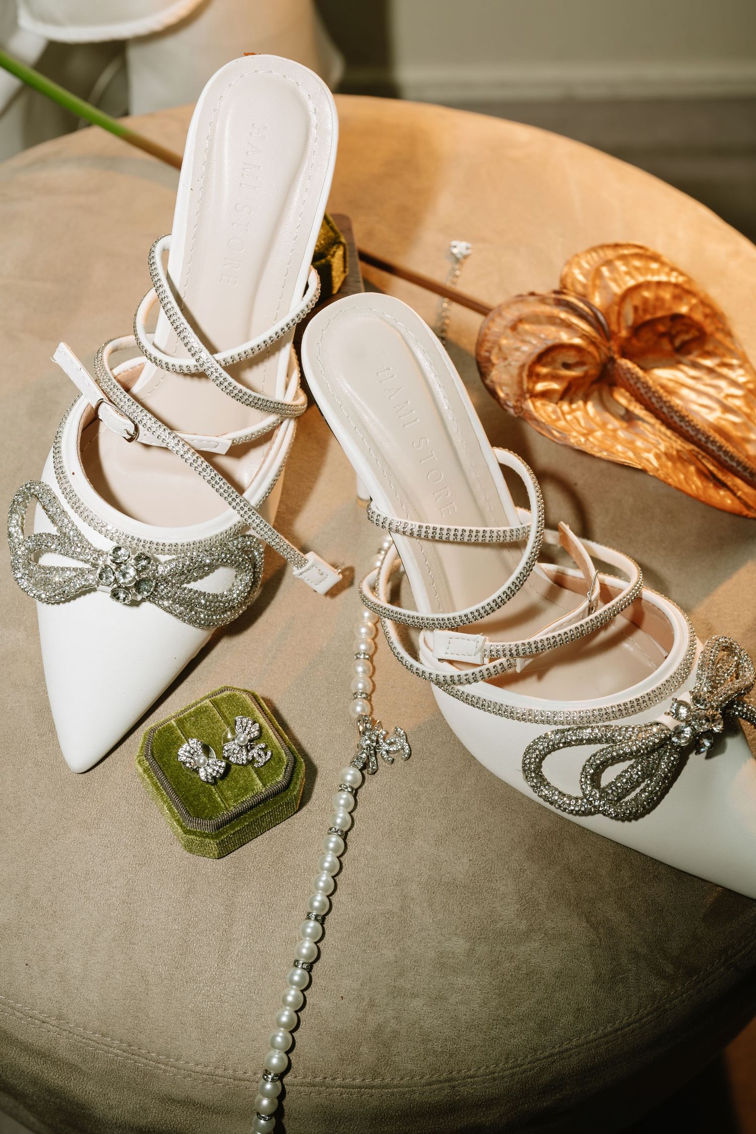 Elegant bridal heels with crystal bow embellishments and strappy design displayed beside pearl jewelry and velvet earring box.