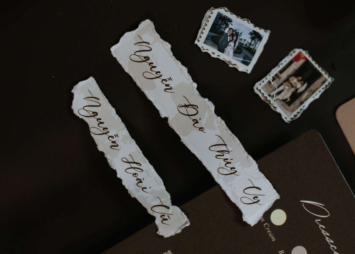 Handwritten calligraphy name tags on torn paper strips beside vintage photo stamps on dark surface.