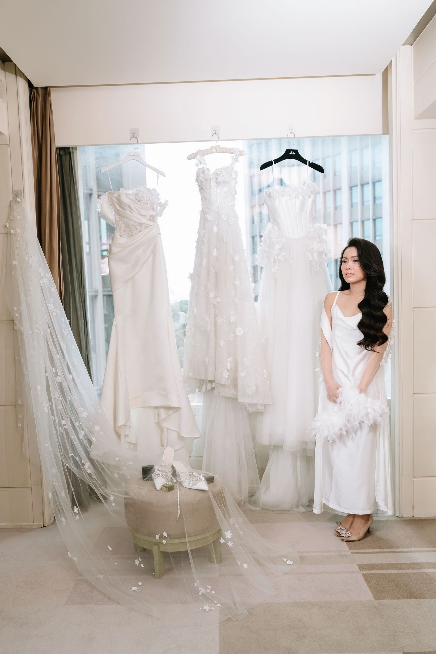 Beautiful bride in white slip dress admiring three elegant wedding gowns hanging by a bright window.