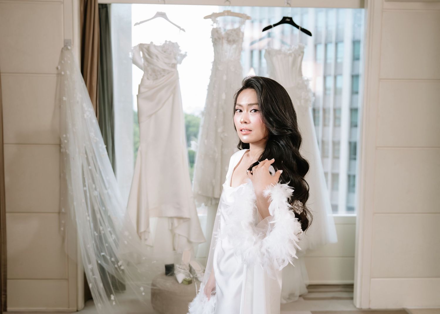Bride in white feather-trimmed robe standing before hanging wedding gowns in bright hotel room.