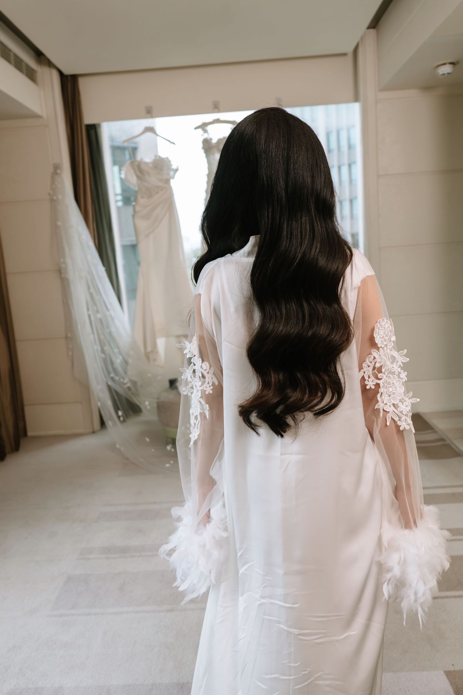 Bride with long wavy dark hair wearing white lace and feather-trimmed bridal robe facing wedding dress.