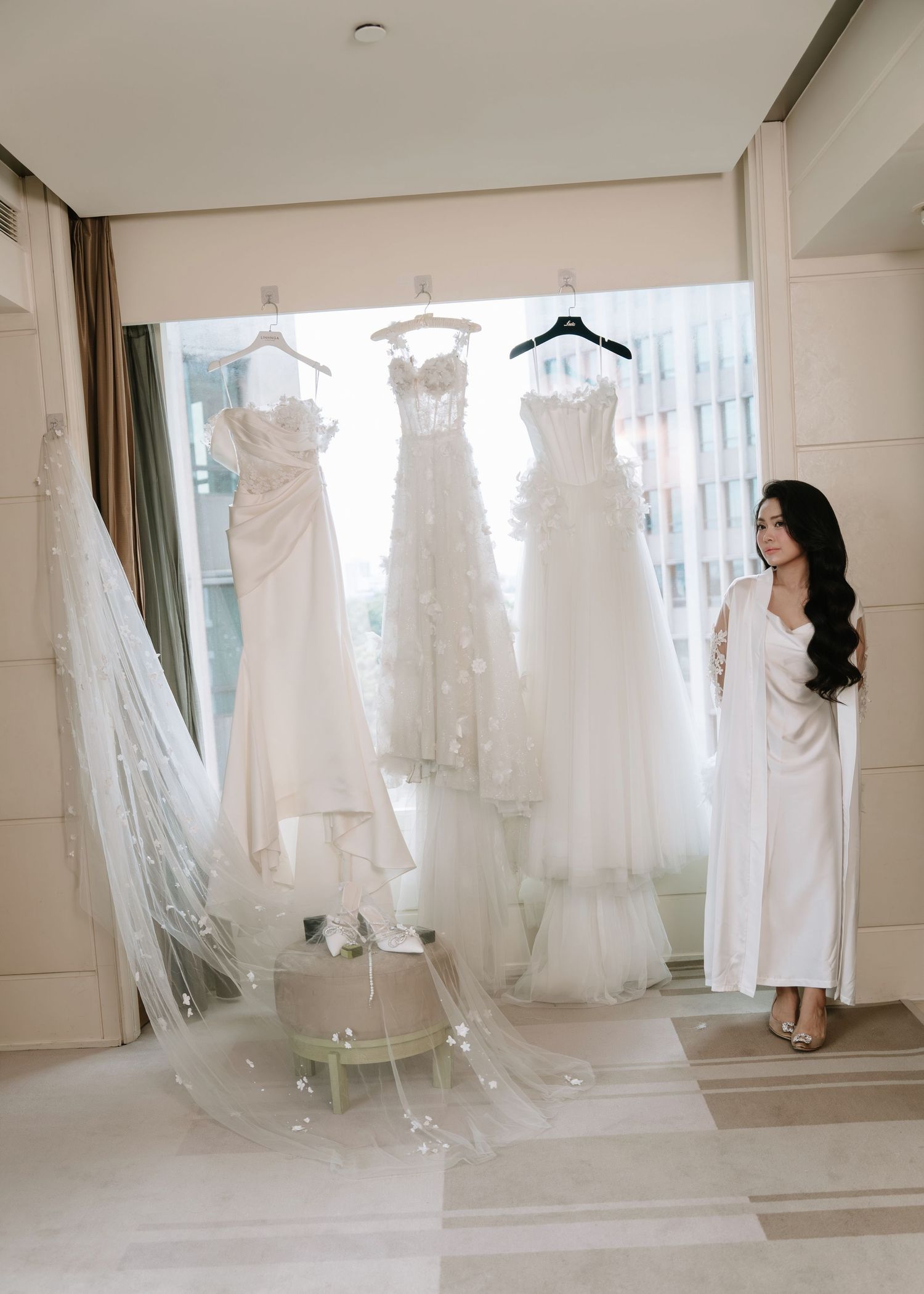 Smiling bride in white robe posing beside three stunning wedding dresses hanging in a sunlit room.
