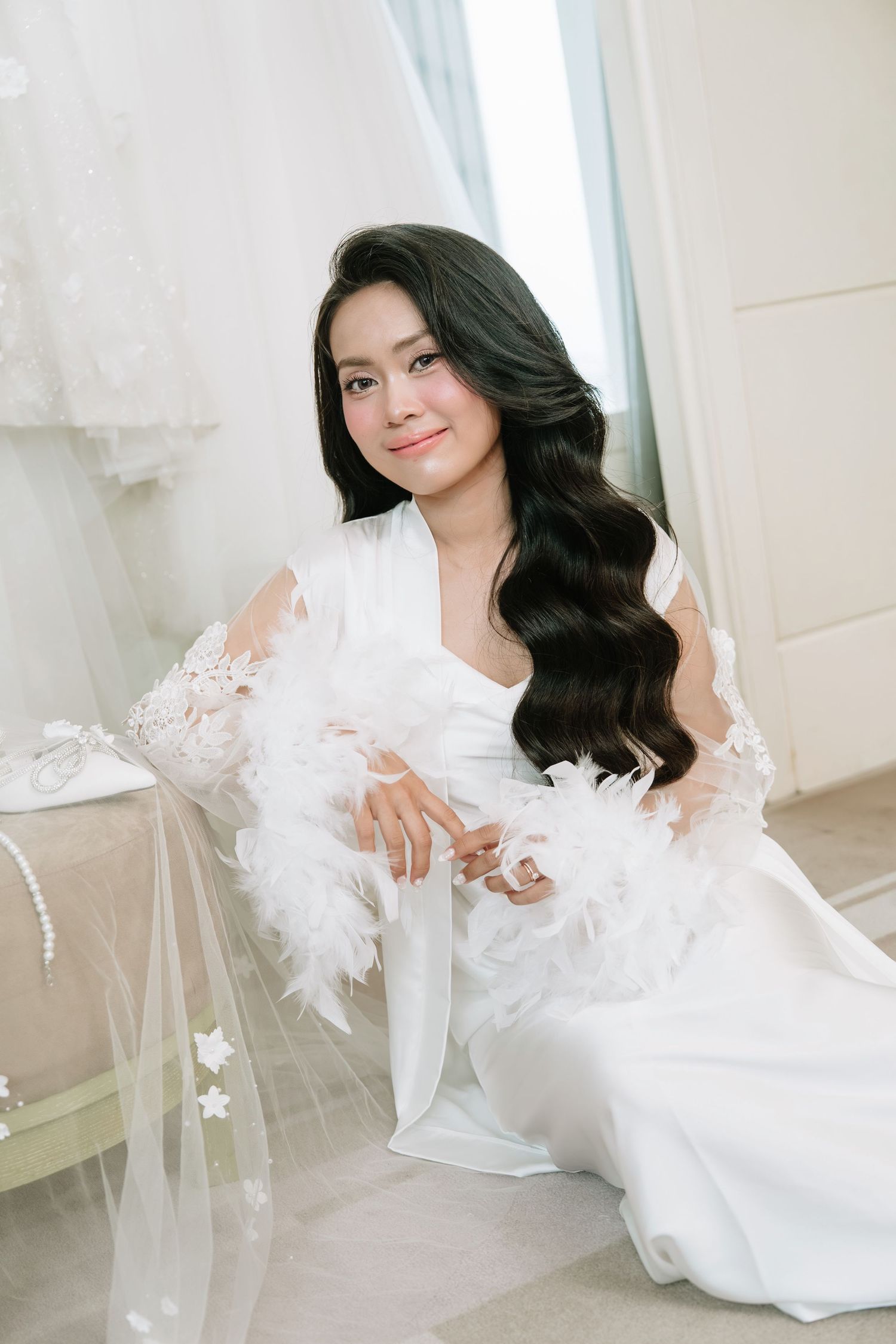 Radiant bride with long wavy hair sitting gracefully in white feather-trimmed robe beside pearl-adorned veil.