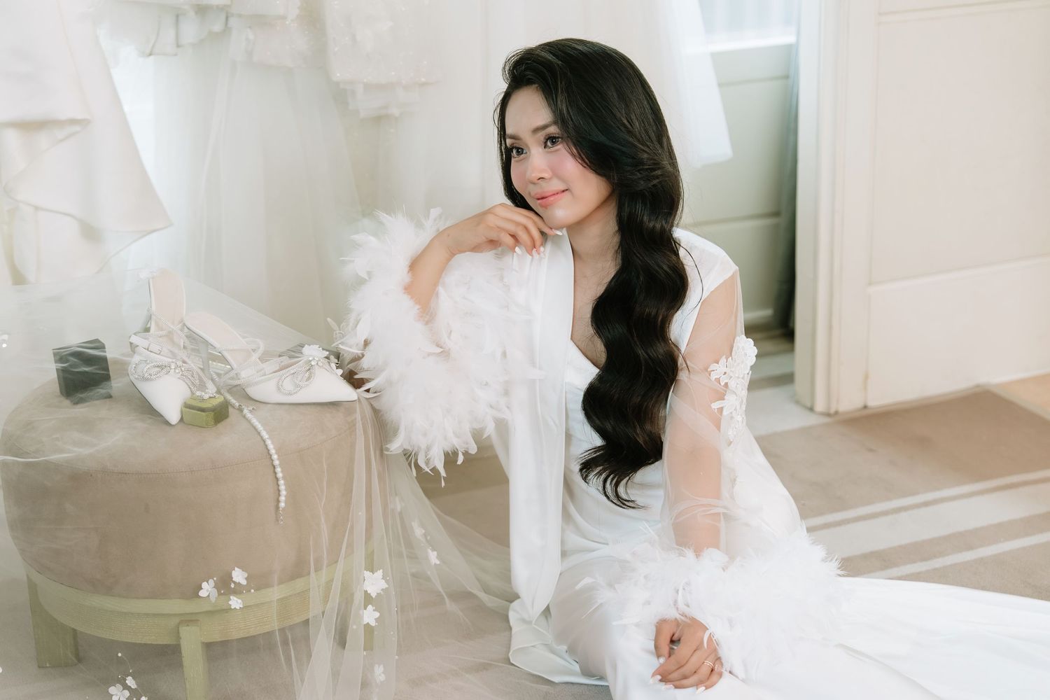 Bride in white feather robe with long wavy hair sitting elegantly in a bridal boutique.