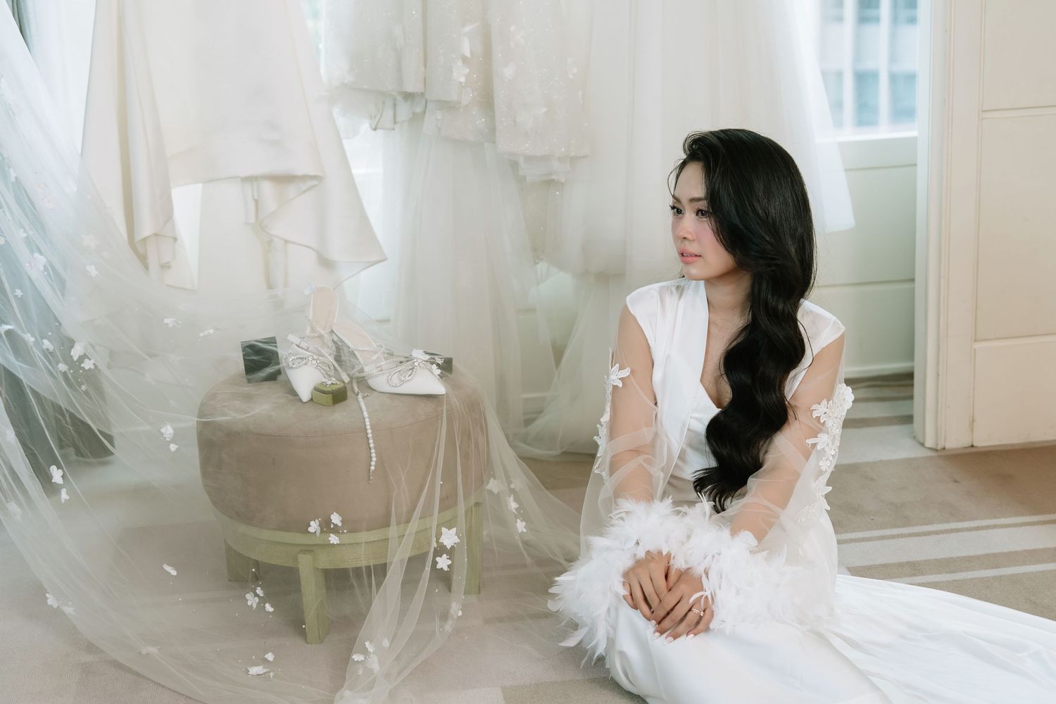 Elegant bride in white gown sitting pensively on floor surrounded by flowing veils in soft light.