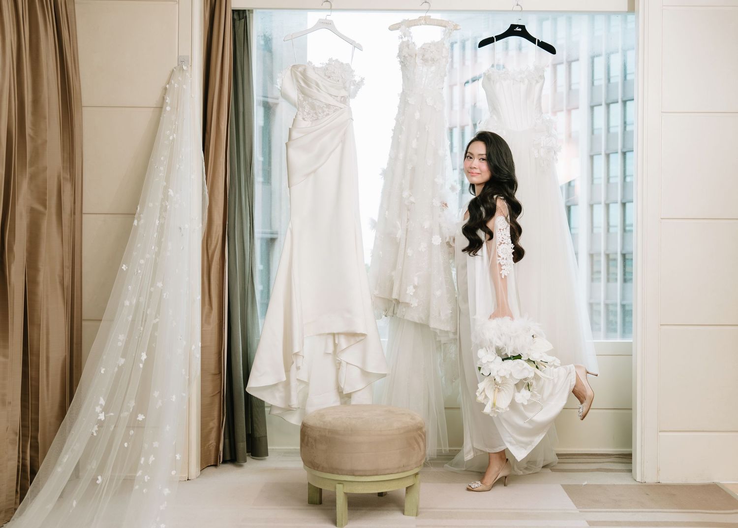 Bride in white gown holding bouquet standing before hanging wedding dresses in bridal suite.