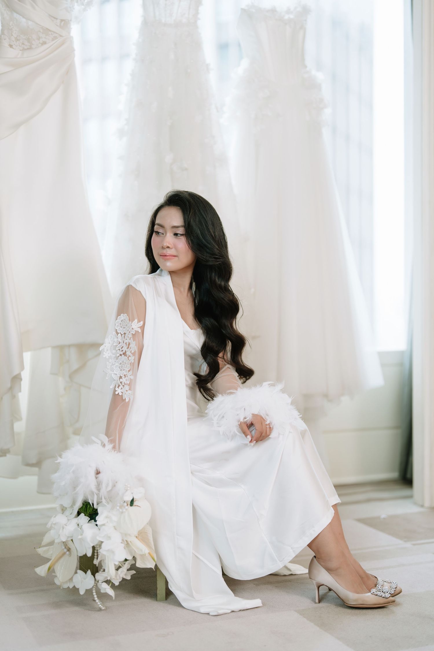 Bride in white lace robe sitting with bouquet surrounded by wedding gowns in bridal shop.