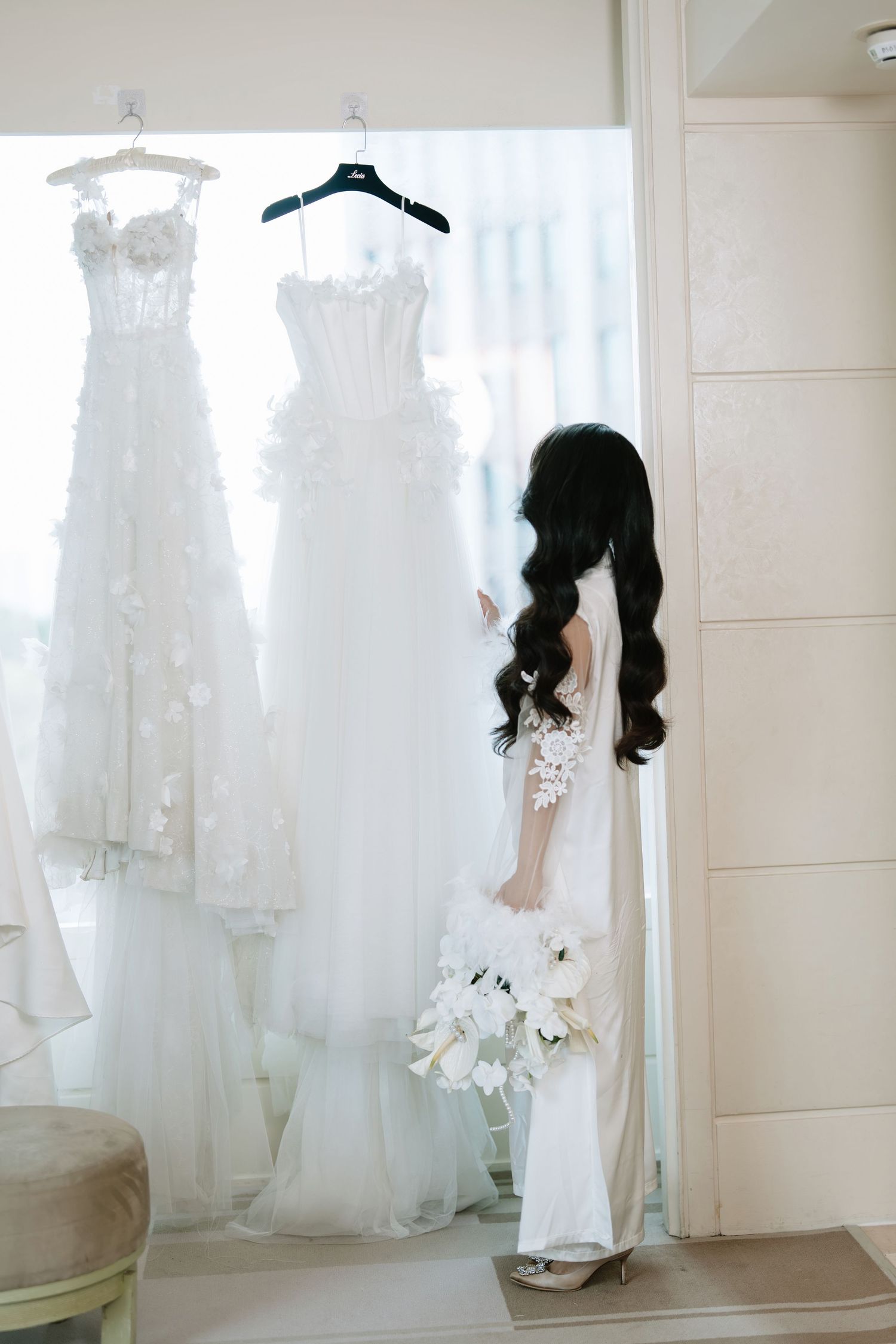 Bride with long dark hair holding bouquet standing before two elegant white wedding gowns.
