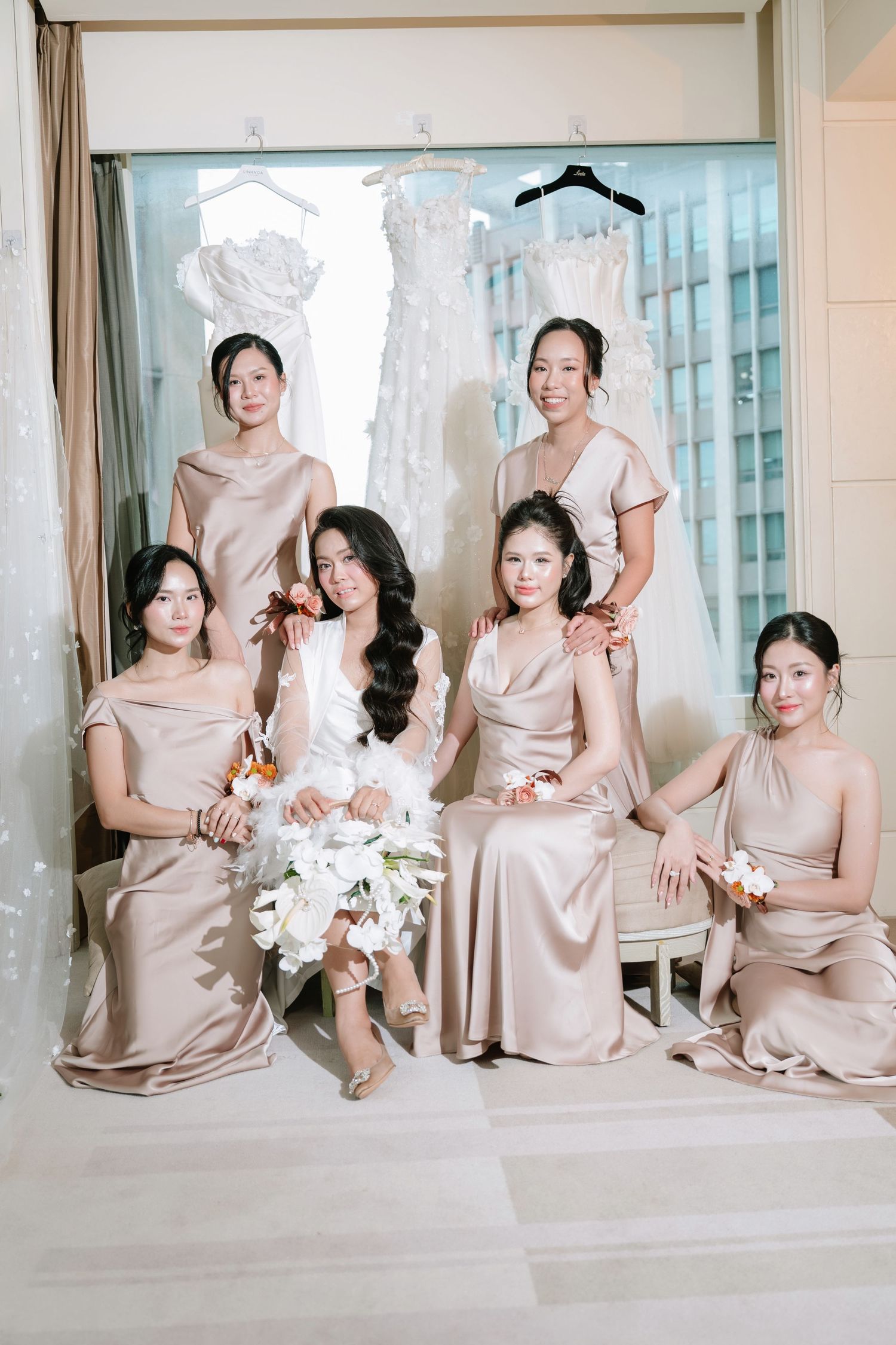 Bride in white robe with five bridesmaids in champagne satin dresses posing in bridal suite.