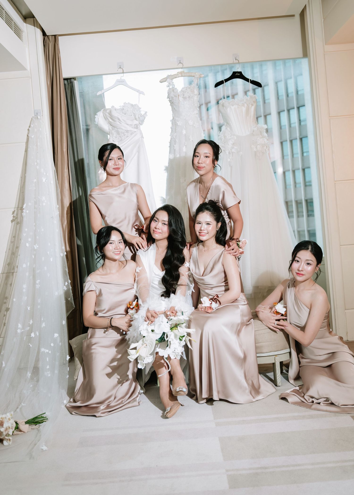 Bride in white holding bouquet posing with five bridesmaids in champagne gowns before wedding dresses.