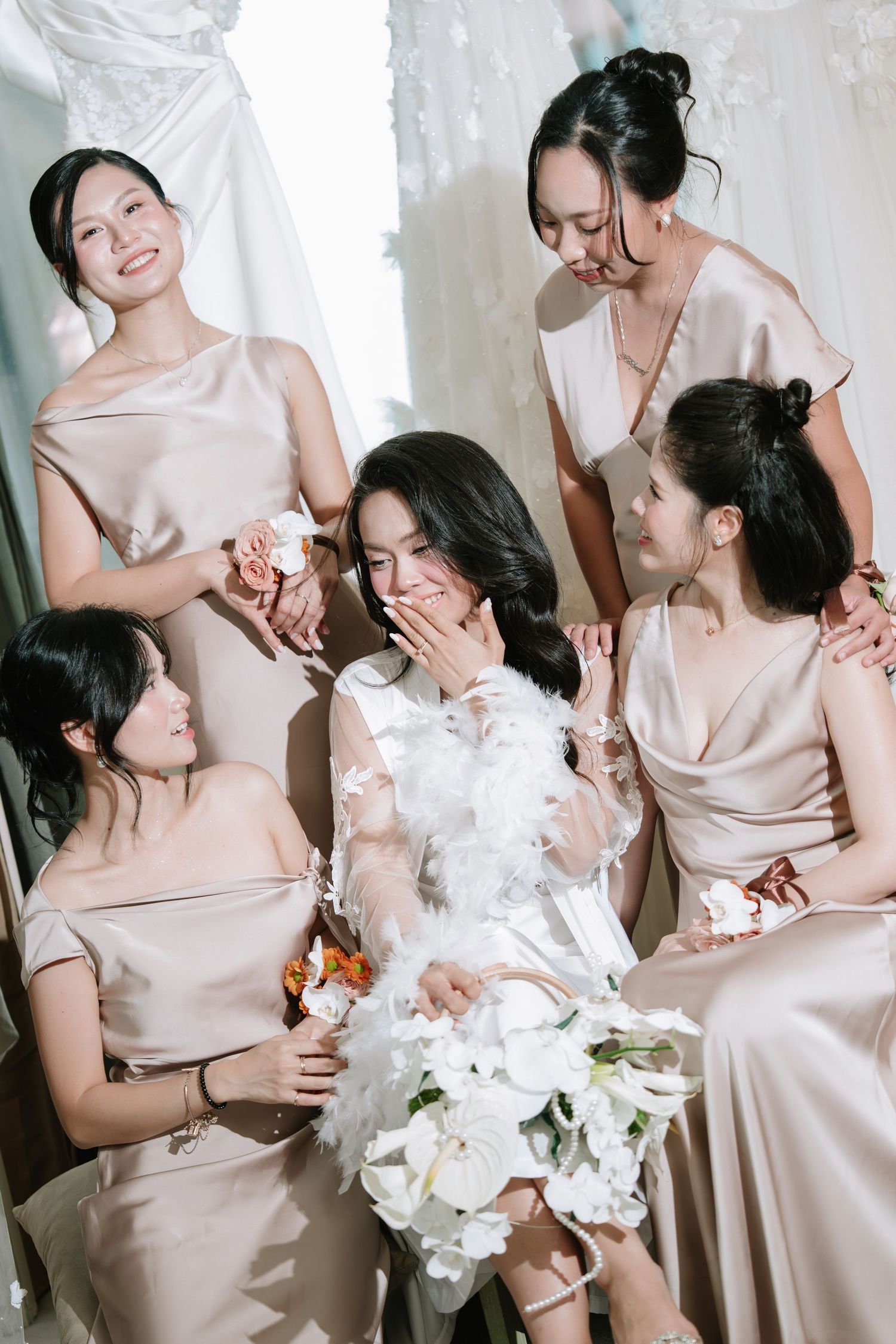 Bride in white feather robe laughing with four bridesmaids in champagne satin dresses holding bouquet.