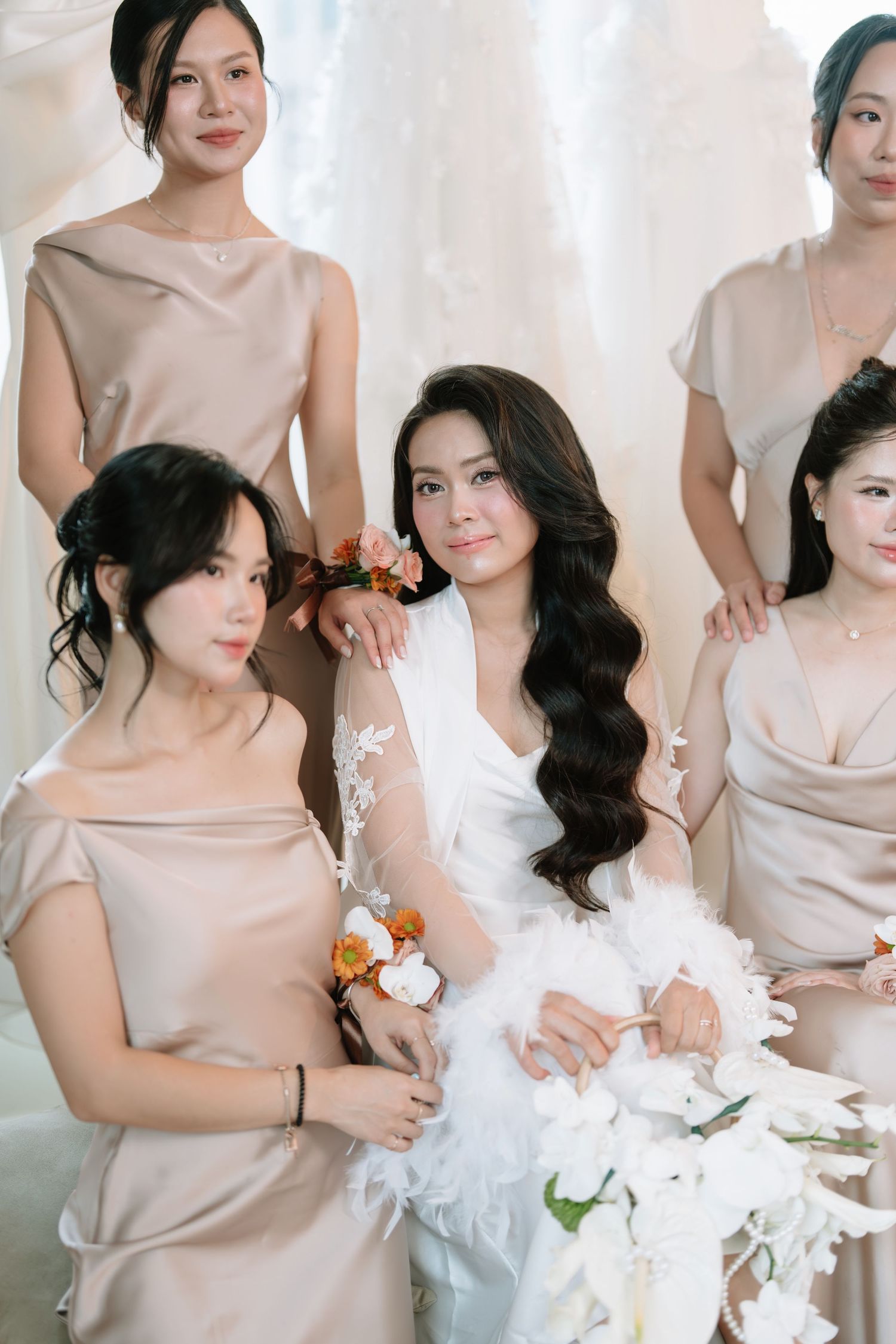 Bride in white feather robe posing with bridesmaids in champagne satin dresses in bridal boutique.