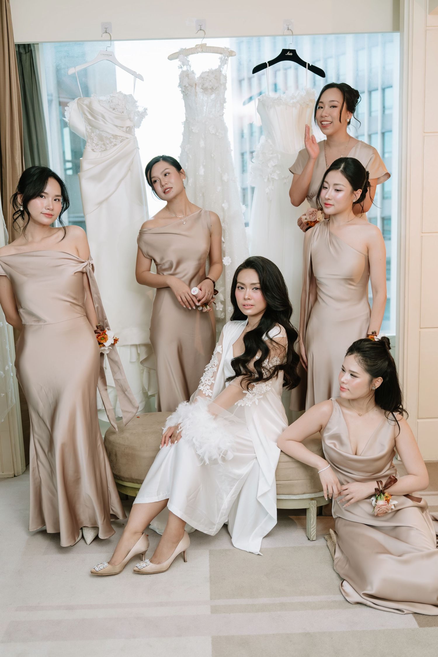Bride in white lace robe seated with five bridesmaids in champagne gowns in elegant bridal suite.