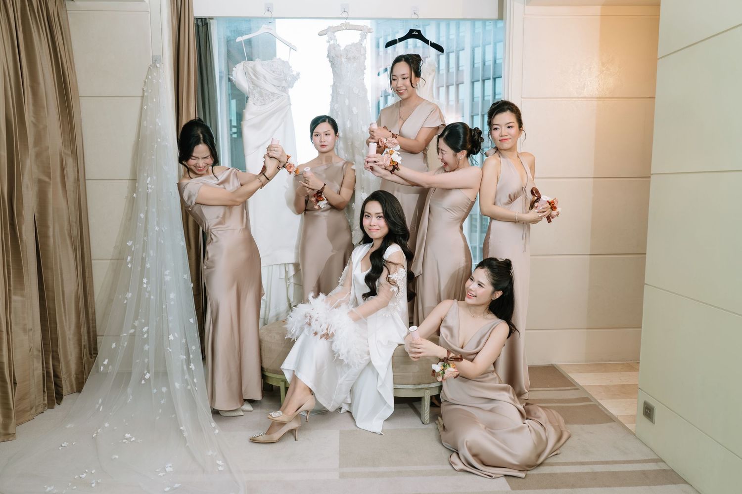 Bride in white gown with veil surrounded by bridesmaids in champagne dresses in bridal suite.