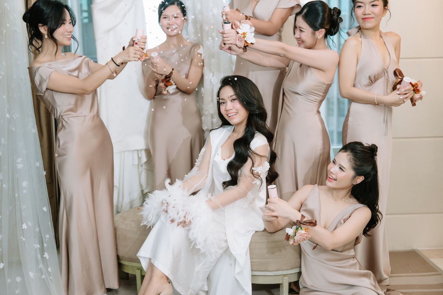 Bride seated on bench surrounded by smiling bridesmaids in champagne satin gowns celebrating with champagne.