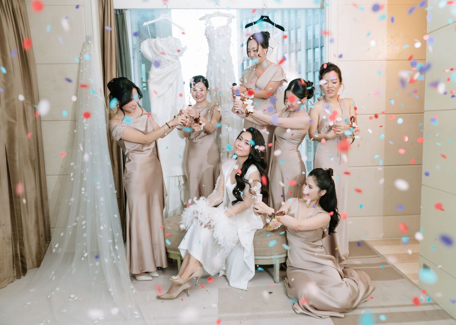 Joyful bride and bridesmaids in nude satin dresses celebrating with colorful confetti in a bridal suite.