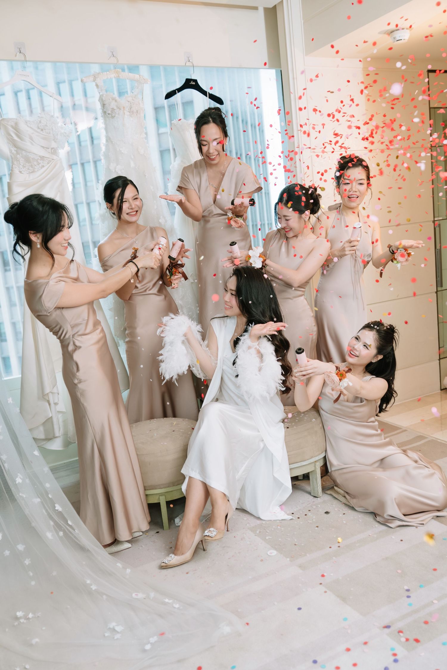 Bride and bridesmaids in champagne gowns laughing as colorful confetti fills the air in a bright room.
