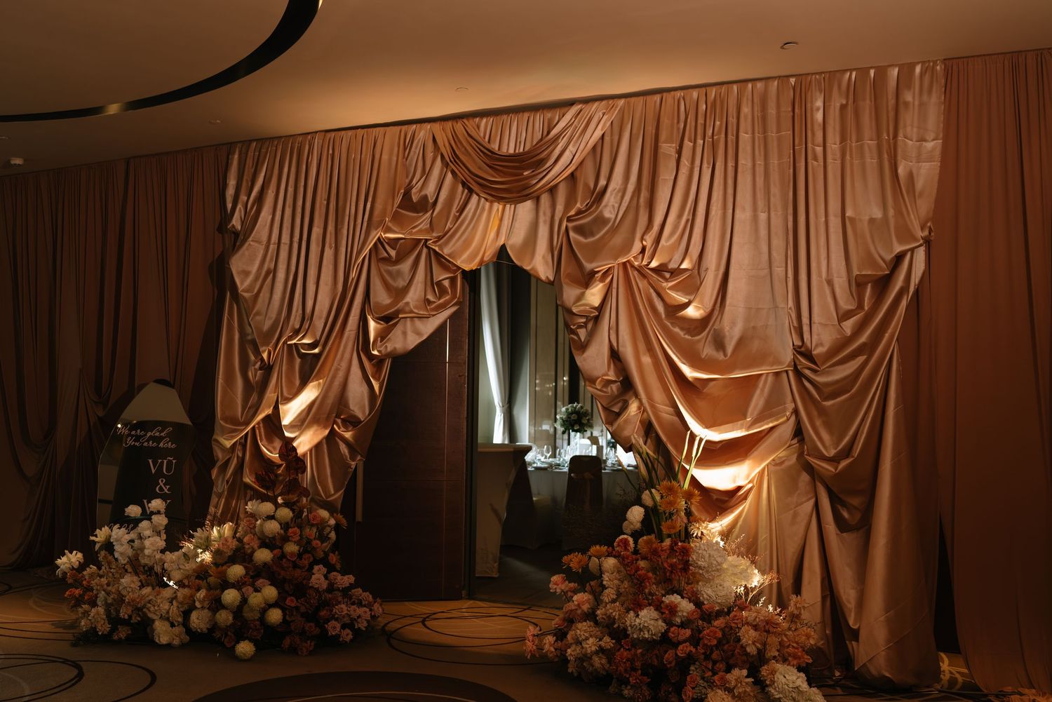 Luxurious champagne satin draped curtain backdrop with soft lighting and floral arrangements for a wedding venue.