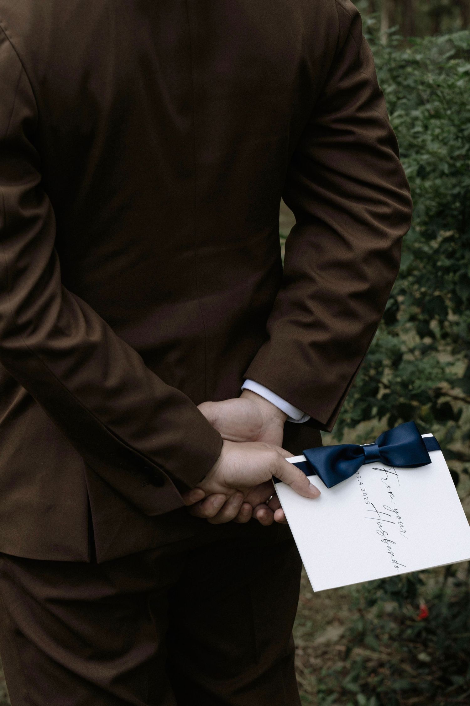 Groom in brown suit holding a blue bow tie and wedding vow card behind his back.