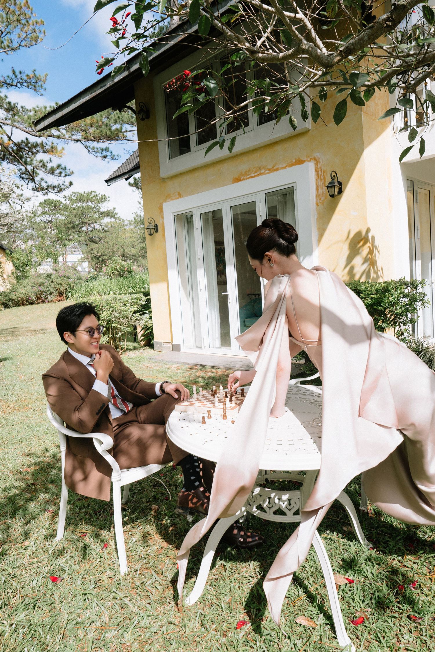 Couple in elegant attire playing chess at white garden table beside a charming yellow cottage.